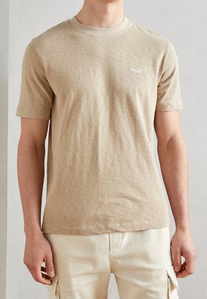 Mann trägt ein beiges kurzärmliges T-Shirt mit einem kleinen weißen "Marc O'Polo Denim"-Logo auf der Brust und hellen Cargohosen.