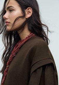 Maglione oversize marrone con colletto a costine; sciarpa rossa a strati con motivi floreali. Capelli scuri lunghi e mossi leggermente visibili.