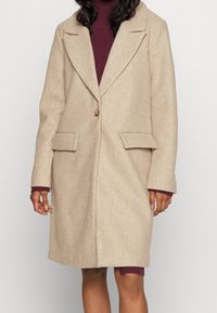 Abrigo de mezcla de lana beige con cuello de pico, cierre de un solo botón y dos bolsillos frontales. Mangas largas con un ajuste estructurado.