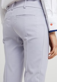 Světle levandulové kalhoty na míru, vyrobené z hladké tkaniny, s úzkým střihem, dvěma zadními kapsami a jemnými detaily v prošívání.