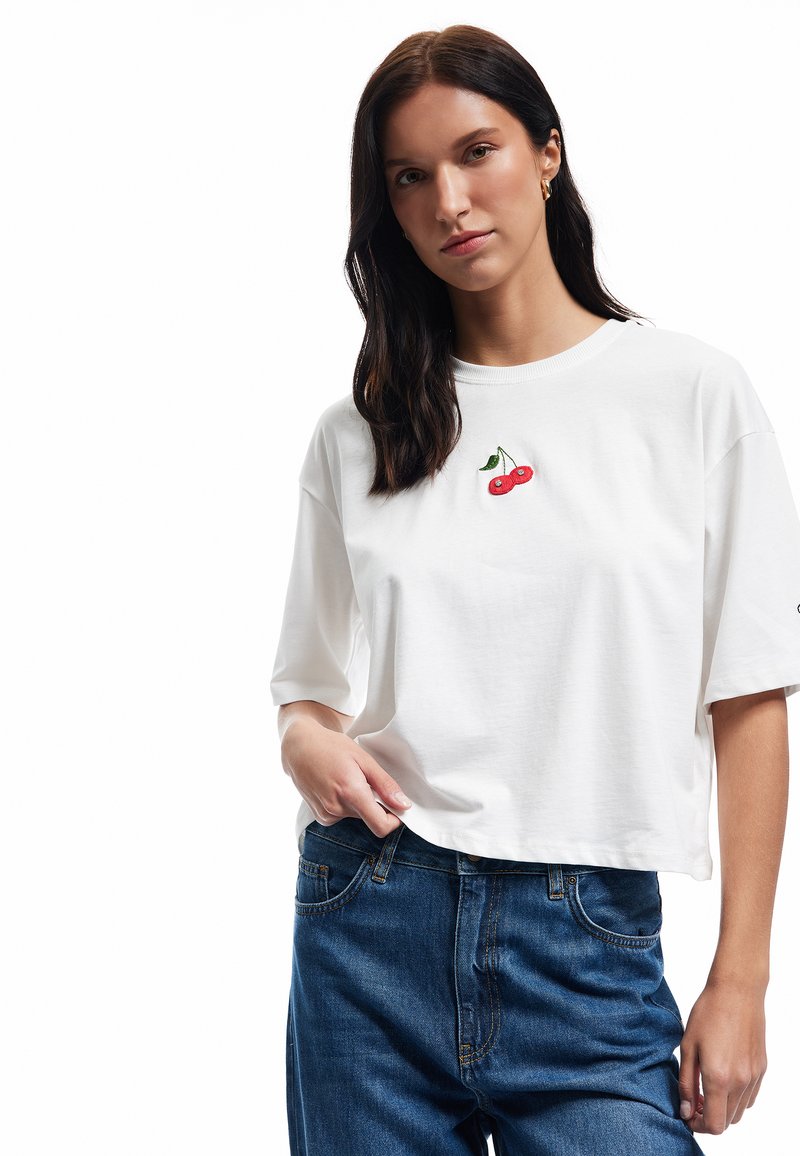 Witte t-shirt met korte mouwen van katoen, voorzien van rode kersen borduursel op de borst, gecombineerd met donkerblauwe denim jeans.