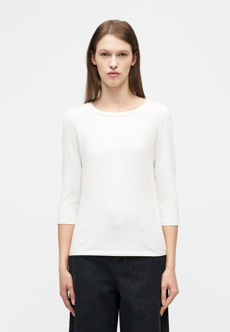 Γυναίκα με ίσια καστανά μαλλιά, ντυμένη με λευκό μπλουζάκι με μανίκια μέχρι τον αγκώνα και μαύρο παντελόνι, να στέκεται μπροστά σε ένα απλό λευκό φόντο.