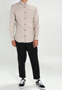 Jasnobeżowa, długa koszula na guziki z kieszenią na piersi, zestawiona z czarnymi, zwężanymi spodniami oraz czarnymi butami sportowymi z białymi akcentami.