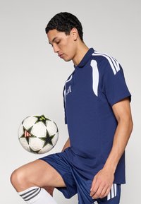 Μπλε σκούρο πόλο μπλουζάκι με άσπρες ρίγες, σε συνδυασμό με μπλε σκούρο σορτς. Το άτομο ισορροπεί μια άσπρη μπάλα ποδοσφαίρου με μαύρα αστέρια.