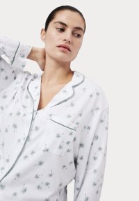 Wit pyjama set met groene paardenbloemprint, voorzien van een zak, kraag en contrasterende groene afwerking langs de randen. Zachte textuur.