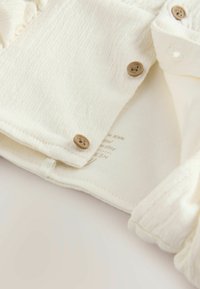 Chemise en coton blanche avec un tissu texturé, dotée de boutons dans une teinte bois naturel et d'une étiquette cousue avec des informations sur la marque à l'intérieur.