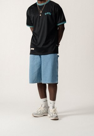 T-shirt en maille noire avec accents turquoise et logo "BLKVIS", associé à un short en jean bleu clair et des baskets gris et blanc.