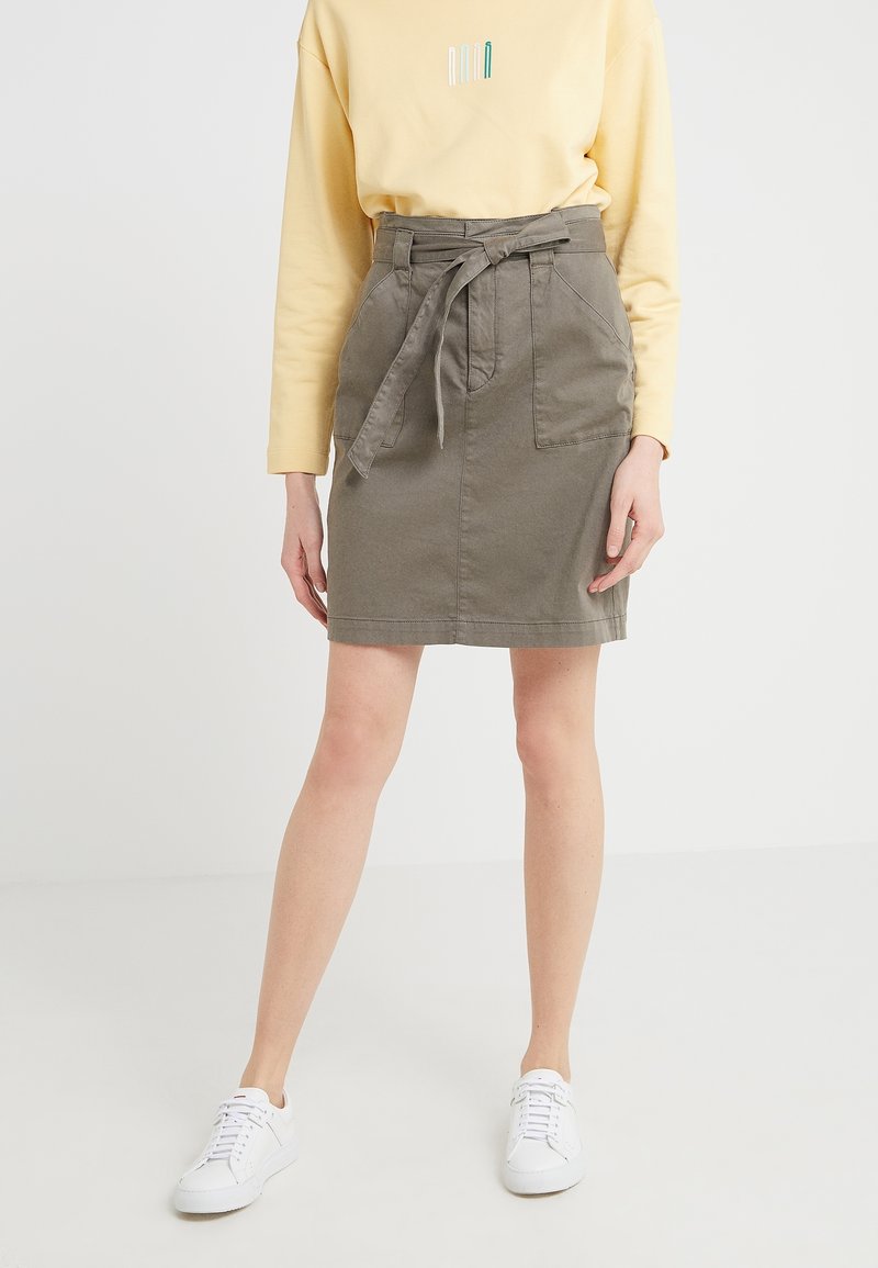 Femme portant un haut beige à manches longues et une jupe taille haute vert olive avec une ceinture nouée et des baskets blanches.