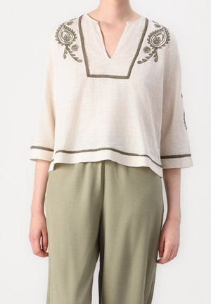 Jasnobeżowa bluzka z oliwkową kwiatową haftem na ramionach i rękawach, zestawiona z luźnymi oliwkowozielonymi spodniami.
