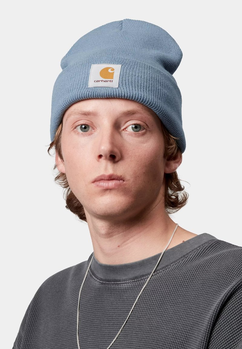 Gorro de punto azul claro con puño doblado y una etiqueta cuadrada amarilla con un logo. Lo lleva una persona con cabello de longitud media.