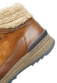 Brązowy zimowy but ze skóry i zamszu z teksturowaną gumową podeszwą oraz beżową polarową wyściółką wokół mankietu przy kostce, widok ukośny.