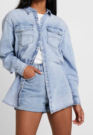 Kvinde iført lys blå denimskjorte med forlommer og matchende denimshorts, hånd i lommen, stående foran en ensfarvet baggrund.