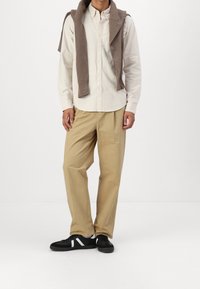 Beige chinos, hvid skjorte med knapper og en brun sweater draperet over skuldrene, parret med sorte sneakers med hvide detaljer.