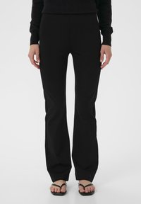 Pantaloni neri svasati con una texture di tessuto liscia, vita alta e dettagli con spacco laterale. Abbinati a sandali neri piatti.