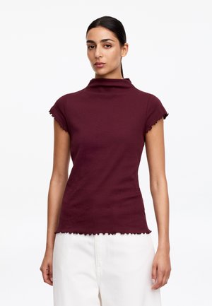 Femme aux cheveux foncés attachés, portant un haut bordeaux à manches courtes à bords volantés et un pantalon blanc taille haute, debout devant un fond blanc.