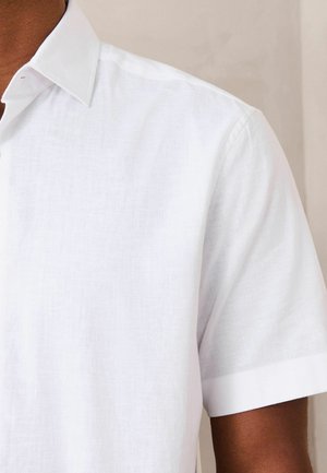 Close-up of a person wearing a white short-sleeve collared shirt against a neutral background.