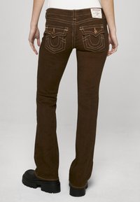 Brązowe dzwony z wyraźnymi tylnymi kieszeniami, kontrastującymi szwami i oznaczoną metką. Gładka faktura i szczegół wytrzymałego materiału.