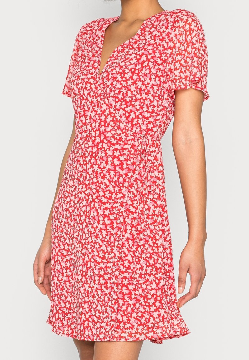 Femme portant une robe portefeuille rouge à manches courtes avec un imprimé floral blanc, debout devant un fond clair uni.