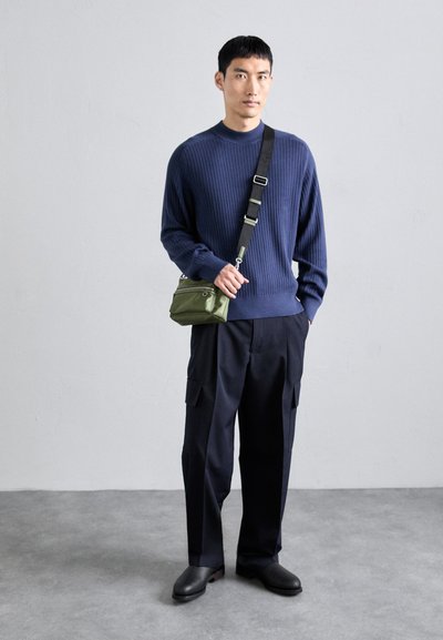 Pulover navy cu model în dungi, pantaloni negri, pantofi negri și geantă crossbody verde. Modelul stă în fața unui fundal neutru, evidențiind designul structurat.