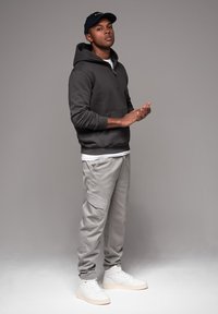 Grijze hoodie, witte t-shirt eronder, lichtgrijze cargo-broek en witte sneakers. Model staat met de armen iets omhoog, met een donkere pet op.