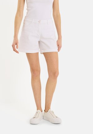 Femme portant un short blanc à ourlet roulé, un débardeur blanc et des baskets blanches à lacets, debout devant un fond clair uni.