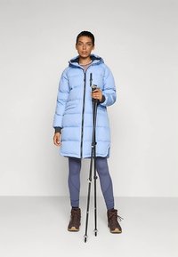 Lichtblauwe donzen jas met een ritssluiting aan de voorkant, gecombineerd met grijze leggings en bruine laarzen. Model houdt wandelstokken vast en staat tegen een neutrale achtergrond.