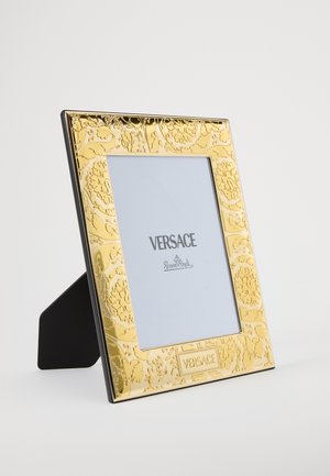 Zelta foto rāmis ar ziedu reljefu un melnu aizmugurējo statīvu. Apakšā redzams "VERSACE" logotips un fotoattēliem paredzēta balta iekšējā matērija.
