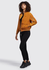 Bomberjacke in leuchtendem Orange mit Reißverschluss, zwei großen Fronttaschen, gerippten Bündchen sowie einem kontrastierenden schwarzen T-Shirt und engen schwarzen Hosen.