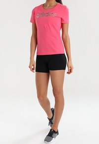 Růžové tričko s krátkým rukávem a reflexním logem "adidas", ve spojení s černými šortkami a šedými sportovními botami, které předvádí přiléhavý střih.