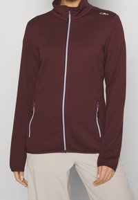 Veste bourgogne avec un col haut, fermeture éclair, et deux poches latérales. Présente un tissu texturé et des détails de fermeture éclair blancs contrastants.