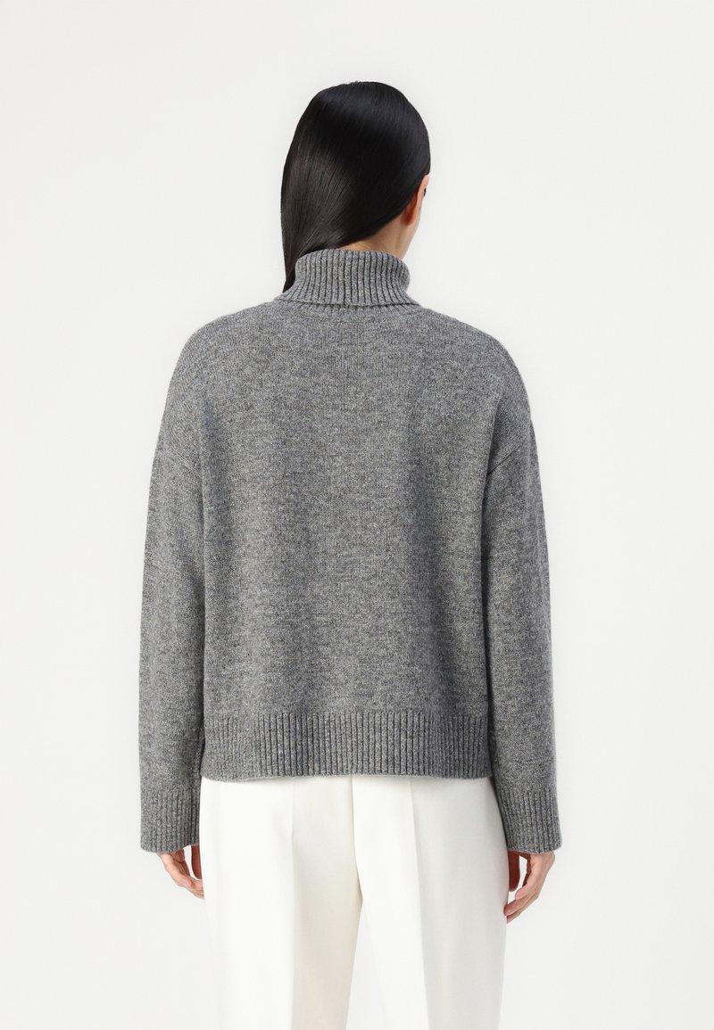 Grauer Rollkragenpullover mit geripptem Saum und Bündchen. Weiches, strukturiertes Strickmaterial. Verkürzte Passform, getragen über weißen Hosen.