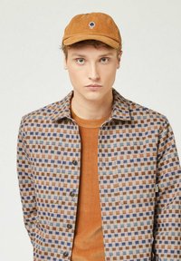 Bruin en blauw geruit overhemd met knopen aan de voorkant, gecombineerd met een oranje ribbelshirt en een corduroy tan pet met een boomlogo.