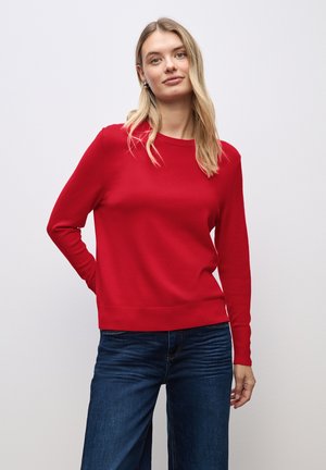 Roter Pullover mit rundem Ausschnitt, langen Ärmeln und gerippten Bündchen. Getragen mit dunkelblauen Jeans. Glatte Textur, schlanker Schnitt, keine sichtbaren Muster.