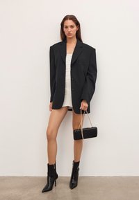 Sieviete, tērpusies pārmērīgā melnā žaketē virs baltas ruched mini kleitas, ar melnām potīšu zābakiem, tur melnu rokassomu ar zelta ķēdi pret vienkāršu sienu.