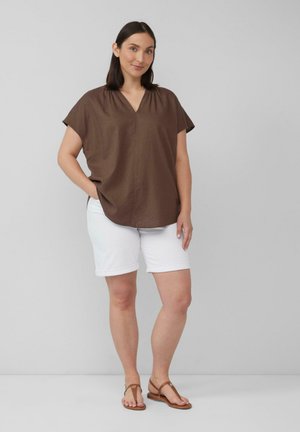 Bruine blouse met korte mouwen en een V-hals, gecombineerd met witte opgerolde shorts. Model draagt bruine sandalen. Eenvoudige, losse pasvorm, gladde textuur.
