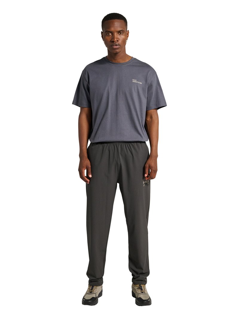 Mand iført mørkegrå løstsiddende t-shirt og sorte afslappede bukser med grå og beige sneakers, stående foran hvid baggrund.