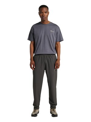 Mann trägt dunkelgraues locker sitzendes T-Shirt und schwarze entspannte Hose mit grauen und beigen Sneakers, steht vor weißem Hintergrund.