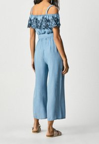Jasnoniebieski dżinsowy kombinezon z dekoltem odkrywającym ramiona, z falbankowym wykończeniem wokół szyi i szerokimi spodniami.
