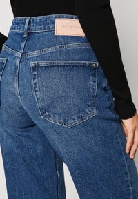 Detailní záběr osoby v modrých džínech s zadními kapsami a béžovou značkovou visačkou Mos Mosh na pase.