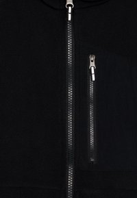 Veste noire zippée présentant une texture en polaire douce, une poche en tissu contrasté avec zip, et des détails de fermeture éclair texturés sur le devant.