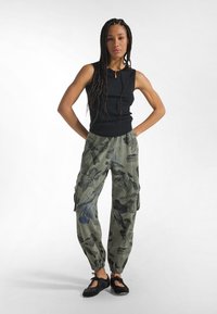 Mujer con el cabello trenzado, de pie con las manos en los bolsillos, llevando una blusa negra sin mangas, pantalones cargo con estampado floral y zapatos planos negros con cordones.