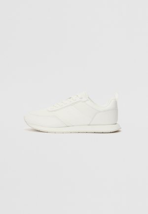 Zapatilla blanca con parte superior de malla y sintético, puntera redonda, cordones planos y una franja lateral texturizada. Suela de goma con un tirador en la parte posterior.