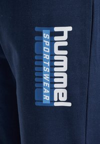 Marineblå sportstoff med en vertikal hvit «hummel»-logo og en blå firkant med «SPORTSWEAR» i hvite bokstaver.