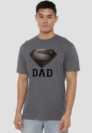 Young man wearing a gray t-shirt with a black metallic Superman logo above the word "DAD," standing against a plain background.