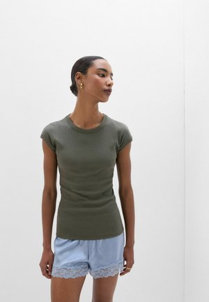 Vrouw met donker haar in een knot draagt een olijfgroene nauwsluitende korte mouw top en lichtblauwe satijnen korte broek met kanten rand, staande tegen een witte muur.