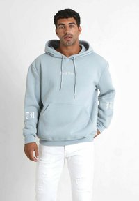 Felpa leggera blu chiaro con tasca a kangaroo frontale, cordini e testo bianco "Sixth June" sul petto. Il manica presenta il testo "TH". Indossata con jeans bianchi strappati.