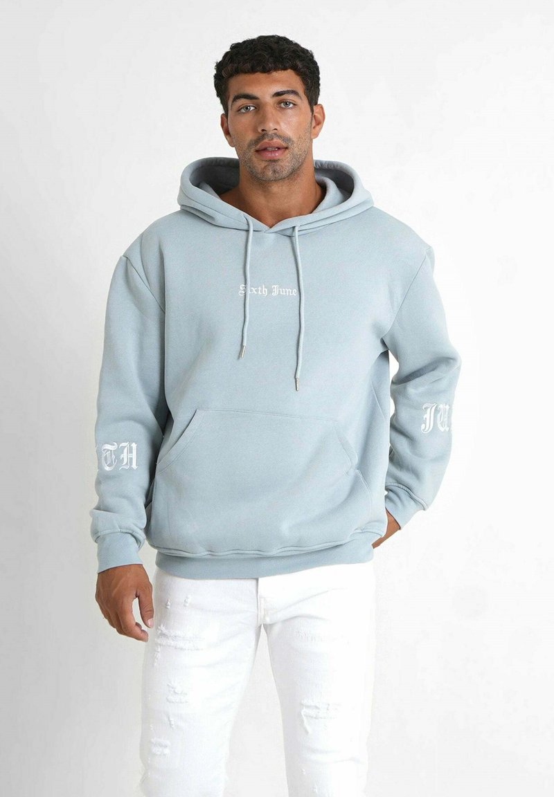 Sudadera de color azul claro con bolsillo canguro frontal, cordones y texto blanco "Sixth June" en el pecho. La manga presenta el texto "TH". Combinada con jeans blancos rasgados.