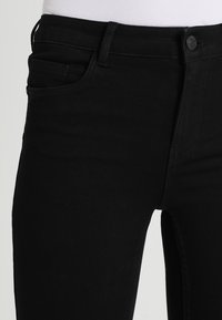 Vaqueros de mezclilla negra con textura suave, diseño de cinco bolsillos, cierre con botones y estilo ajustado, con detalles de costuras visibles.