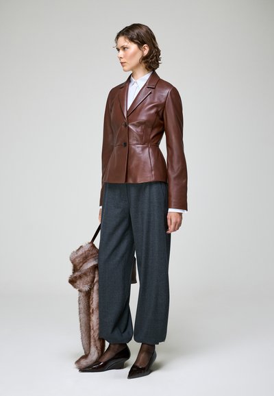 Femme portant un blazer en cuir marron, un pantalon gris à jambes larges, des chaussures foncées et tenant un manteau en fourrure sur un fond clair uni.