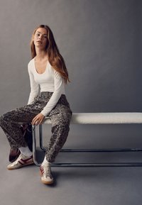 Weißes Langarmshirt, lockere Hose mit Leopardenmuster, beige Sneaker mit roten Akzenten, sitzend auf einer weißen, strukturierten Bank vor einem grauen Hintergrund.